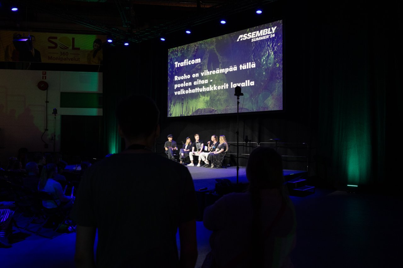 Panelistit lavalla, kuva otettu kaukaa yleisön takaa, joten myös keskustelijoiden takana oleva valkokangas näkyy. Valkokankaalla lukee paneelin nimi: 