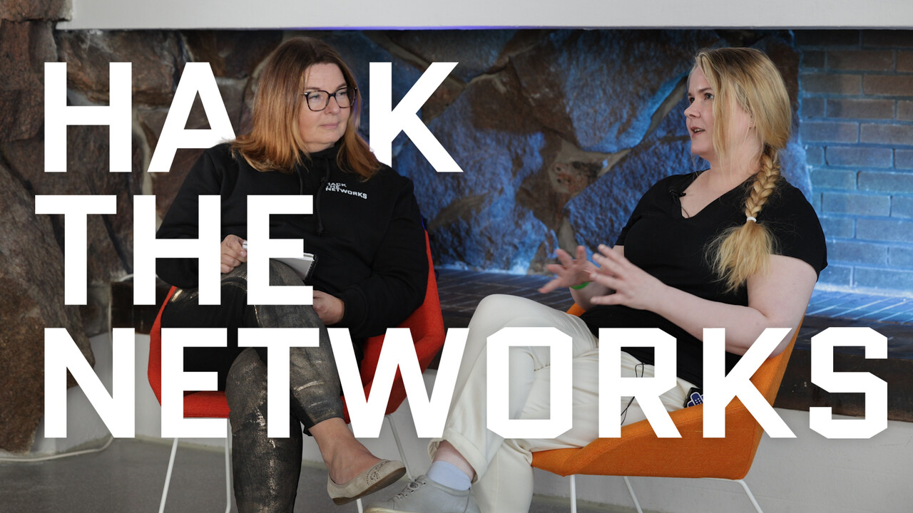 Teknologia- ja strategiajohtaja Kirsi Karlamaa haastattelemassa valkohattuhakkeri Mariana Leppäahoa, kumpikin istuu nojatuolissa takan edustalla. Hack the Networksin logo on editoitu ympäröimään heitä. 