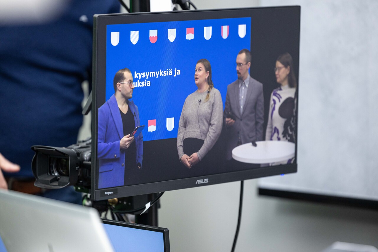 valokuva videokameran monitorista: videokameran näytöllä neljä asiantuntijaa keskustelemassa, taustalla kirkkaansininen powerpont-grafiikka, jossa on abstrakteja vaakunoiden kuvia. 